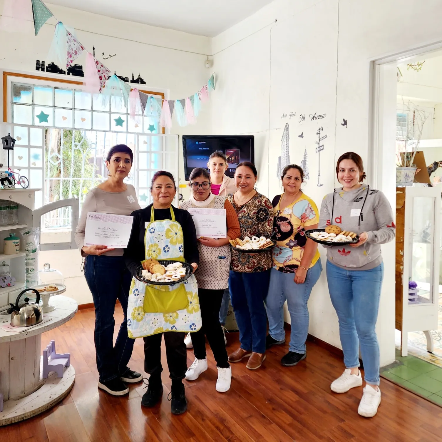 Grupo de mujeres en un taller de galletas, cada una sosteniendo su diploma.
